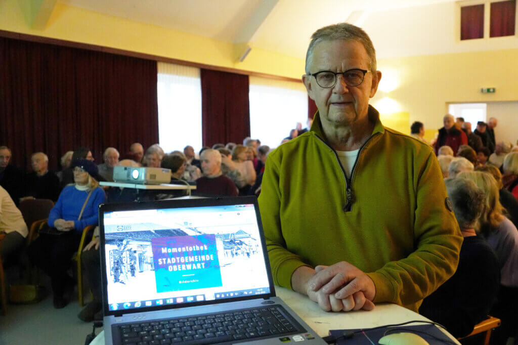 Tillfried Schober, Betreiber der Momentothek, steht an einem Tisch mit offenem Laptop auf dem die Startseite der Webseite geöffnet ist.