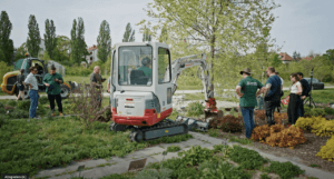 Ein kleiner Bagger steht in einem Garten. Rundherum sind Menschen, die bei der Arbeit zusehen