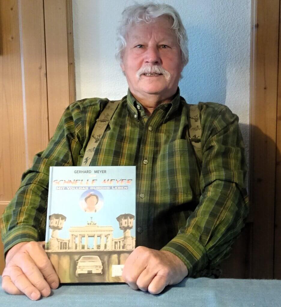 Auf dem Bild seiht man Gerhard Meyer. Er trägt ein grünes kariertes Hemd und hält sein Buch mit dem Titel "Der schnelle Meyer mit Vollgas durchs Leben.