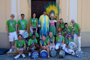 Eine Gruppe von etwa fünfzehn Personen posiert fröhlich vor einem Gebäude. Die meisten tragen grüne T-Shirts mit weißen Hosen oder Shorts. Ein Mann in der Mitte trägt ein farbenfrohes Gewand, und eine Frau daneben trägt ein Dirndl mit einem auffälligen, gelben und blauen Kopfschmuck. Mehrere Mitglieder der Gruppe halten Trommeln oder andere Musikinstrumente.