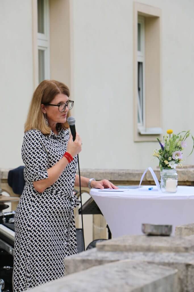 Heidi Adelwöhrer, Präsidentin der IV-Burgenland, steht an einem weißen Stehtisch und hält ein Mikrofon in der Hand. Sie trägt ein gemustertes Kleid und eine Brille. Auf dem Tisch befinden sich ein Blatt Papier, ein blauer Kugelschreiber, und ein Glasgefäß mit einem kleinen Blumenstrauß und einer weißen Kerze. Im Hintergrund ist eine helle Gebäudewand mit zwei Fenstern zu sehen.