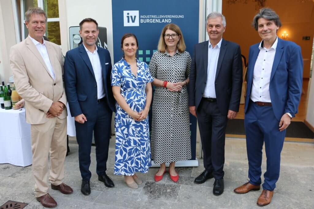 Christian Strasser, Matthias Unger, Christina Glocknitzer, Heidi Adelwöhrer, Johann Pinterits und Johannes Ernst stehen nebeneinander vor einem Banner mit der Aufschrift "Industriellenvereinigung Burgenland." Christian Strasser trägt einen beigen Anzug mit weißem Hemd. Matthias Unger ist in einen dunkelblauen Anzug mit weißem Hemd gekleidet. Christina Glocknitzer trägt ein blau-weiß gemustertes Kleid. Heidi Adelwöhrer ist in ein schwarz-weiß gemustertes Kleid und rote Schuhe gekleidet. Johann Pinterits trägt einen dunkelgrauen Anzug mit weißem Hemd. Johannes Ernst ist in einen blauen Anzug mit weißem Hemd und braunen Schuhen gekleidet. Im Hintergrund sind ein geöffnetes Fenster und eine geöffnete Tür zu sehen, die zu einem Raum mit Kronleuchtern führen.