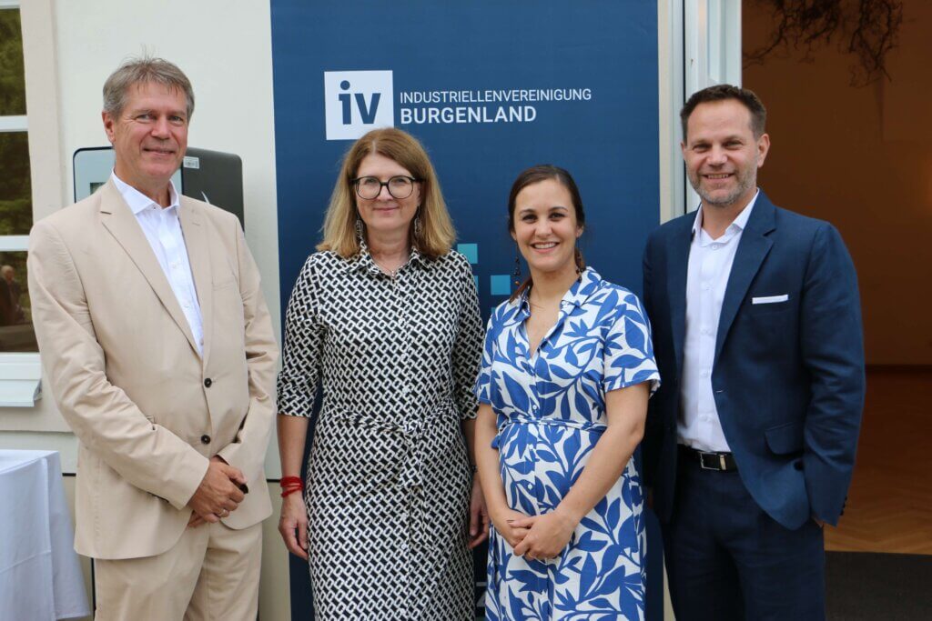 Das IV Burgenland Präsidium posiert vor einem Werbebanner der Industriellenvereinigung Burgenland. Von links nach rechts sind zu sehen: IV-Burgenland Vizepräsident Christian Strasser (PET to PET Recycling Austria GmbH) in einem beigen Anzug, IV-Burgenland Präsidentin Heidi Adelwöhrer (Neudoerfler Office Systems GmbH) in einem gemusterten Kleid, IV-Burgenland Vizepräsidentin Christina Glocknitzer (Seal Maker Produktions- und Vertriebs GmbH) in einem blauen und weißen gemusterten Kleid und IV-Burgenland Vizepräsident Matthias Unger (Unger Steel) in einem dunkelblauen Anzug.