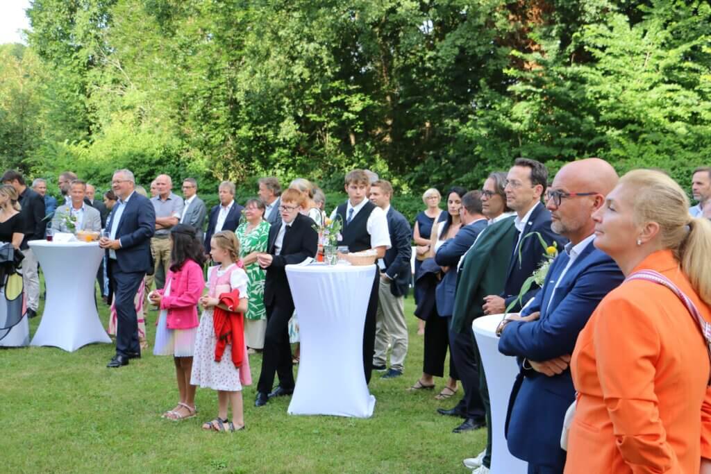 Eine Gruppe von etwa dreißig Personen steht auf einer grünen Wiese, umgeben von dichtem, grünem Laubwerk. Die Gäste, die unterschiedliche elegante Kleidung tragen, unterhalten sich in kleinen Gruppen und stehen teilweise an hohen, weißen Stehtischen mit Blumenarrangements. Im Vordergrund tragen zwei kleine Mädchen bunte Kleider, eines in Weiß und Rosa mit einer roten Jacke, das andere in Weiß mit einem rosa Blazer. Rechts vorne ist eine Frau in einem orangefarbenen Blazer zu sehen. Die Atmosphäre ist entspannt und gesellig, die Gäste lauschen aufmerksam oder unterhalten sich.