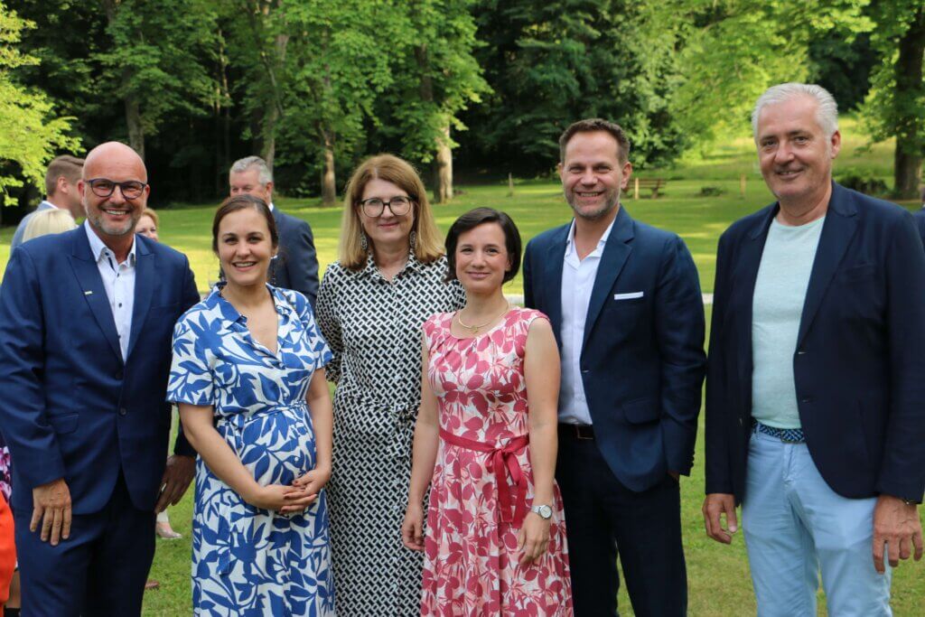 Martin Geiger, Christina Glocknitzer, Heidi Adelwöhrer, Aniko Benkö, Matthias Unger und Manfred Gerger stehen nebeneinander auf einer Wiese und lächeln in die Kamera. Martin Geiger trägt einen blauen Anzug und eine Brille. Christina Glocknitzer ist in einem blau-weißen Kleid mit floralem Muster gekleidet. Heidi Adelwöhrer trägt ein schwarz-weiß gemustertes Kleid. Aniko Benkö ist in ein pink-weiß gemustertes Kleid gekleidet. Matthias Unger trägt einen blauen Anzug ohne Krawatte, und Manfred Gerger ist in einem dunkelblauen Sakko mit hellblauem Hemd und hellblauen Hosen gekleidet. Im Hintergrund ist eine grüne Parklandschaft zu sehen.