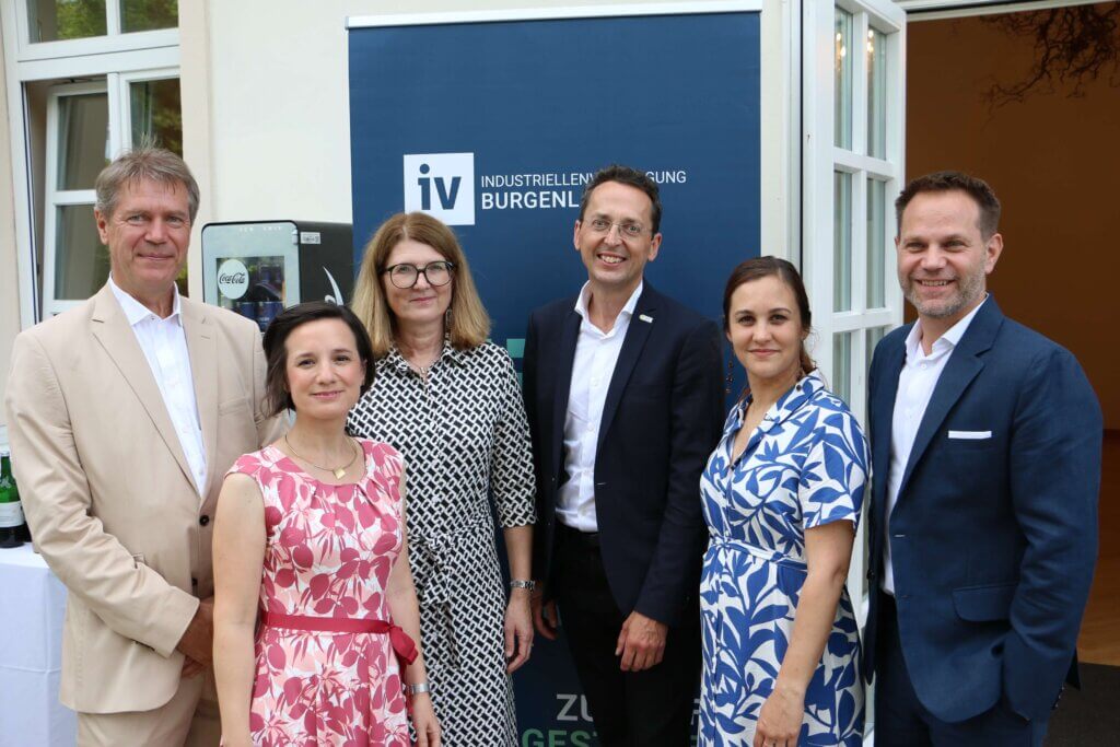 Christian Strasser, Aniko Benkö, Heidi Adelwöhrer, Bernhard Puttinger, Christina Glocknitzer und Matthias Unger stehen nebeneinander vor einem Banner mit der Aufschrift "Industriellenvereinigung Burgenland." Christian Strasser trägt einen beigen Anzug mit weißem Hemd. Aniko Benkö ist in ein pink-weiß gemustertes Kleid gekleidet. Heidi Adelwöhrer trägt ein schwarz-weiß gemustertes Kleid und eine Brille. Bernhard Puttinger trägt einen dunkelblauen Anzug mit weißem Hemd und keine Krawatte. Christina Glocknitzer trägt ein blau-weiß gemustertes Kleid. Matthias Unger ist in einen dunkelblauen Anzug mit weißem Hemd gekleidet. Im Hintergrund ist ein offenes Fenster zu sehen.