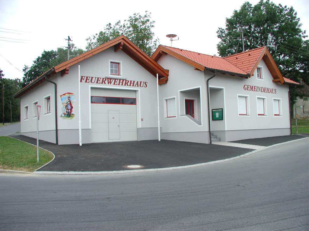 Ein modernes Gebäude mit der Aufschrift „Feuerwehrhaus“ und „Gemeindehaus“. Das Gebäude hat weiße Wände und ein rotes Dach. Auf der linken Seite des Gebäudes ist ein farbenfrohes Wandbild zu sehen.