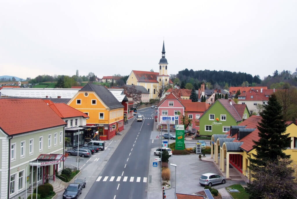 Eine lebendige Straßenszene von Kaindorf, mit bunten Gebäuden auf beiden Seiten der Straße. In der Mitte steht eine Kirche mit einem hohen Turm und einem Dach aus roten Ziegeln, umgeben von orangefarbenen, gelben und grünen Gebäuden. Auf der Straße sind parkende Autos, ein paar Fußgänger und ein klarer Himmel darüber zu sehen. Die Stadt liegt eingebettet in eine grüne Landschaft mit einer Mischung aus Feldern und Bäumen im Hintergrund.