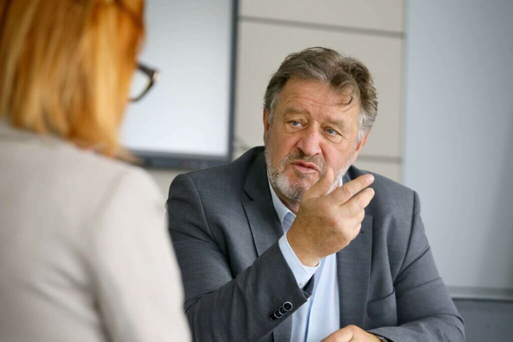 Bildungsdirektor Alfred Lehner in einem Gespräch. Er trägt einen grauen Anzug und ein hellblaues Hemd. Mit ernster Miene und lebhafter Gestik spricht er mit einer Frau, die mit dem Rücken zur Kamera sitzt. Sie hat schulterlanges, blondes Haar und trägt eine Brille. Im Hintergrund sind unscharfe Büroelemente zu sehen.