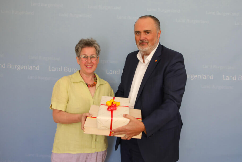 Regina Petrik und Landeshauptmann Hans Peter Doskozil stehen nebeneinander und halten gemeinsam ein Geschenk, das in helles Papier mit rotem und gelbem Band verpackt ist. Regina Petrik trägt eine helle grüne Bluse und eine bunte Halskette, während Hans Peter Doskozil einen dunkelblauen Anzug mit weißem Hemd trägt. Beide lächeln. Im Hintergrund ist eine hellblaue Wand mit der Aufschrift „Land Burgenland“ mehrfach zu sehen.