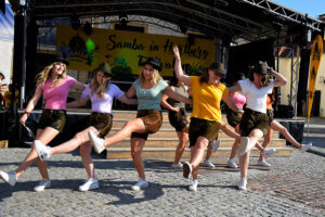 Eine Gruppe von Frauen führt einen Tanz auf der Bühne des Samba-Festivals in Hartberg auf. Sie tragen bunte T-Shirts, Lederhosen und Hüte. Die Bühne ist mit einem großen Banner des Festivals im Hintergrund dekoriert.