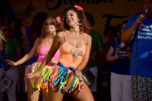 Zwei Frauen tanzen auf der Bühne des Samba-Festivals. Sie tragen bunte, fröhliche Kostüme mit Glitzer und Blumendekorationen im Haar. Weitere Mitglieder der Sambagruppe sind im Hintergrund zu sehen, ebenfalls in farbenfroher Kleidung.