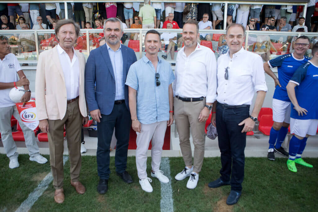 Kurt Maczek, Heinrich Dorner, Robert Hergovich, Mario Windhofer und Leonhard Schneemann stehen nebeneinander auf einem Fußballplatz. Im Hintergrund sind Zuschauer auf einer Tribüne zu sehen. Einige Zuschauer stehen, während andere sitzen und den Blick auf die Kamera richten. Links steht ein Mann in weißem Poloshirt mit BFV-Logo, rechts zwei Männer in blauen Fußballtrikots.