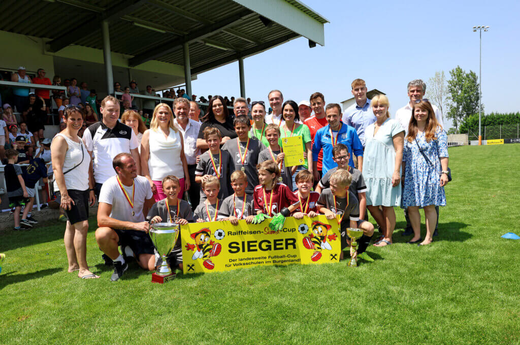 Die Mannschaft der Volksschule Draßburg posiert auf einem Fußballfeld. Die Jungen tragen graue Trikots und Medaillen, einige halten Pokale und eine Urkunde. Vor ihnen ist ein gelbes Banner mit der Aufschrift "Raiffeisen-Sumsi-Cup SIEGER". Im Hintergrund stehen Bildungslandesrätin Daniela Winkler (Mitte, in weißem Oberteil), Bildungsdirektor Burgenland Alfred Lehner (links neben Daniela Winkler) und andere Erwachsene, die lächeln. Auf der überdachten Tribüne im Hintergrund sind zahlreiche Zuschauer zu sehen.