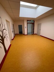 Ein weiterer Flur im neuen Kindergarten, ebenfalls mit hellen Wänden und Baummotiven. Der gelbe Boden und die roten Fußleisten sind auch hier zu sehen. Am Ende des Flurs befindet sich eine Glastür, die in einen weiteren Bereich des Kindergartens führt. Ein Oberlicht sorgt für viel natürliches Licht.