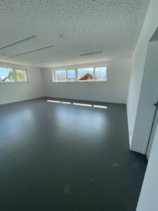 Ein großer Raum im neuen Kindergarten Loipersdorf-Kitzladen. Die Wände sind weiß gestrichen und der Boden ist dunkelgrau. Die Decke ist ebenfalls weiß und hat kleine Löcher, die für eine gute Akustik sorgen. Durch die breiten Fenster auf zwei Seiten des Raumes fällt viel Tageslicht herein, wodurch der Raum hell und einladend wirkt. Im Hintergrund sind Bäume und ein Teil eines Hauses zu sehen.