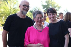 Michel Reimon, Regina Petrik und Sigi Maurer stehen zusammen und lächeln. Michel Reimon trägt ein schwarzes T-Shirt und eine Brille. Regina Petrik trägt eine leuchtend pinke Bluse und eine bunte Perlenkette, sie hält ein Glas in der Hand. Sigi Maurer trägt ein schwarzes Kleid und lächelt ebenfalls. Im Hintergrund sind verschwommen weitere Personen zu sehen, die sich im Freien unterhalten.