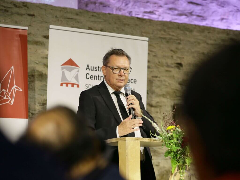 Norbert Darabos, Präsident des Austrian Centre for Peace, hält eine Begrüßungsrede. Er steht an einem Rednerpult, hält ein Mikrofon und trägt einen schwarzen Anzug mit weißem Hemd und Krawatte. Vor ihm steht ein Blumengesteck. Im Hintergrund sind ein Banner des Austrian Centre for Peace und eine Steinwand zu sehen. Die Atmosphäre ist feierlich und professionell. Foto: LEXI