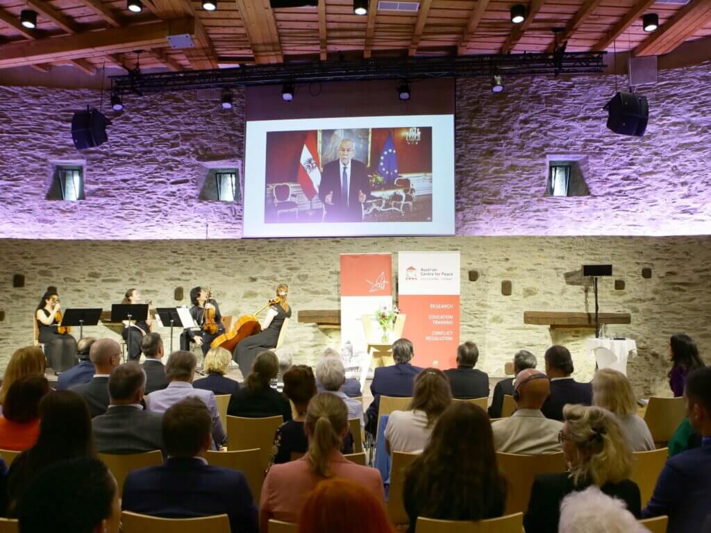 Das Foto zeigt eine Videobotschaft von Bundespräsident Alexander Van der Bellen, die während des Forums abgespielt wurde. Auf einer Leinwand ist er zu sehen, wie er vor den Flaggen Österreichs und der Europäischen Union spricht. Im Vordergrund spielt ein Streichquartett, was die festliche und bedeutungsvolle Stimmung der Veranstaltung weiter hervorhebt. Das Publikum verfolgt aufmerksam die Botschaft des Bundespräsidenten. Foto: Lexi