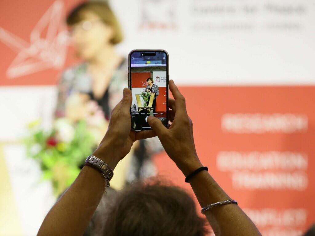 Hände einer Person halten ein Smartphone, das ein Foto von Andrea Eberl, einer Rednerin auf dem 2. Österreichischen Forum für Frieden auf Burg Schlaining, aufnimmt. Andrea Eberl steht unscharf im Hintergrund vor einer roten Wand mit weißen Schriftzügen und einem Origami-Kranich-Logo. Im Vordergrund ist ein Blumenarrangement leicht verschwommen zu sehen. Foto: Lexi