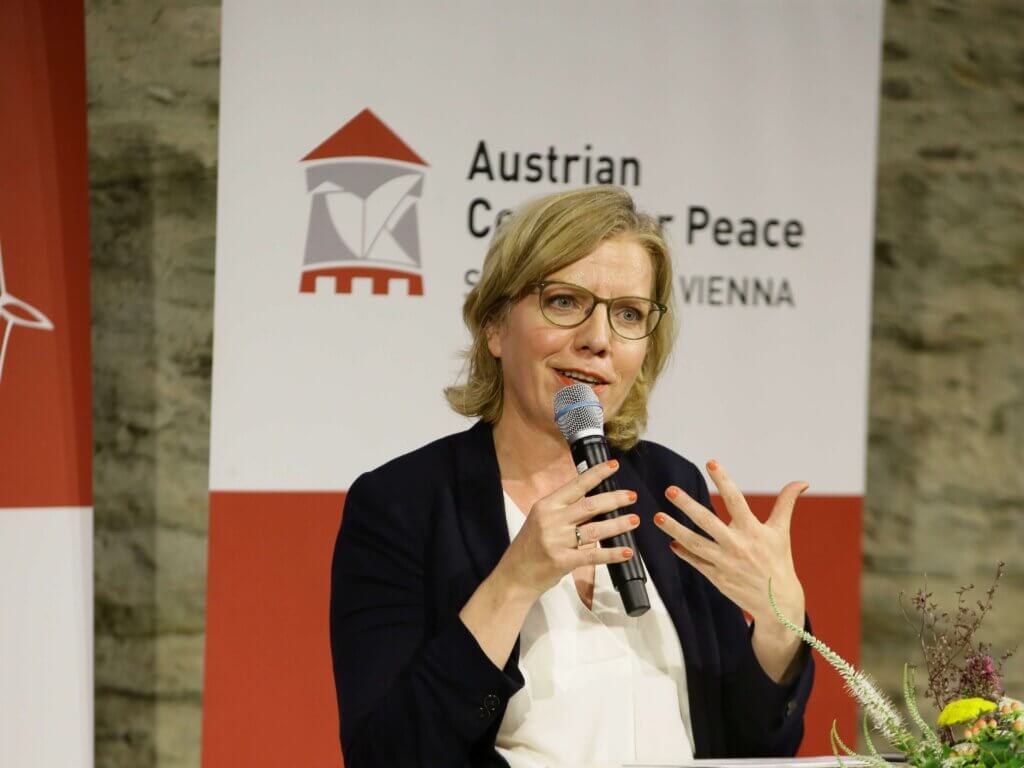 Leonore Gewessler, Bundesministerin für Klimaschutz, Umwelt, Energie, Mobilität, Innovation und Technologie, spricht bei der 2. Austrian Forum for Peace auf Burg Schlaining. Sie hält ein Mikrofon in der rechten Hand und gestikuliert mit der linken. Sie trägt eine Brille, eine weiße Bluse und ein dunkles Sakko. Im Hintergrund sind ein Banner mit der Aufschrift "Austrian Center for Peace" und eine gemauerte Wand zu sehen. Rechts unten im Bild befinden sich einige Blumen. Foto: Lexi