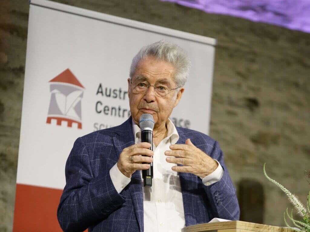 Dr. Heinz Fischer, ehemaliger österreichischer Bundespräsident, hält eine Ansprache beim 2. Austrian Forum for Peace auf Burg Schlaining. Er trägt ein weißes Hemd und ein blau kariertes Sakko. In der rechten Hand hält er ein Mikrofon, die linke Hand ist erhoben. Im Hintergrund befindet sich ein Roll-Up mit dem Logo und dem Schriftzug des Austrian Centre for Peace vor einer Steinwand. Foto: Lexi