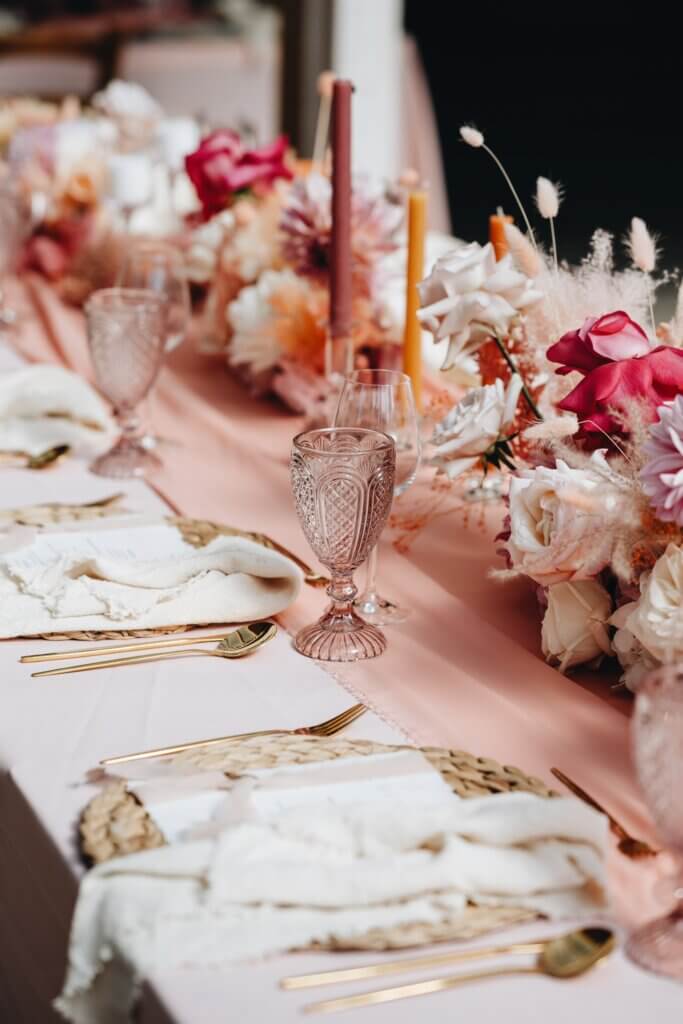 Elegante Tischdekoration mit Kristallgläsern, goldenen Bestecken und Blumenarrangement auf rosa Tischdecke.